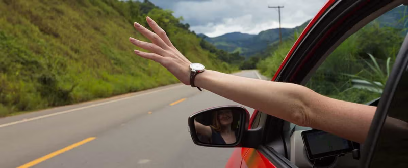 Um braço para fora da janela de um carro em movimento numa estrada rural.
