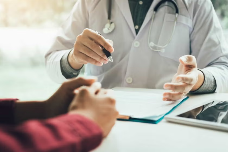 Médico de bata branca a discutir um relatório com um paciente, mostrando um documento numa pasta.